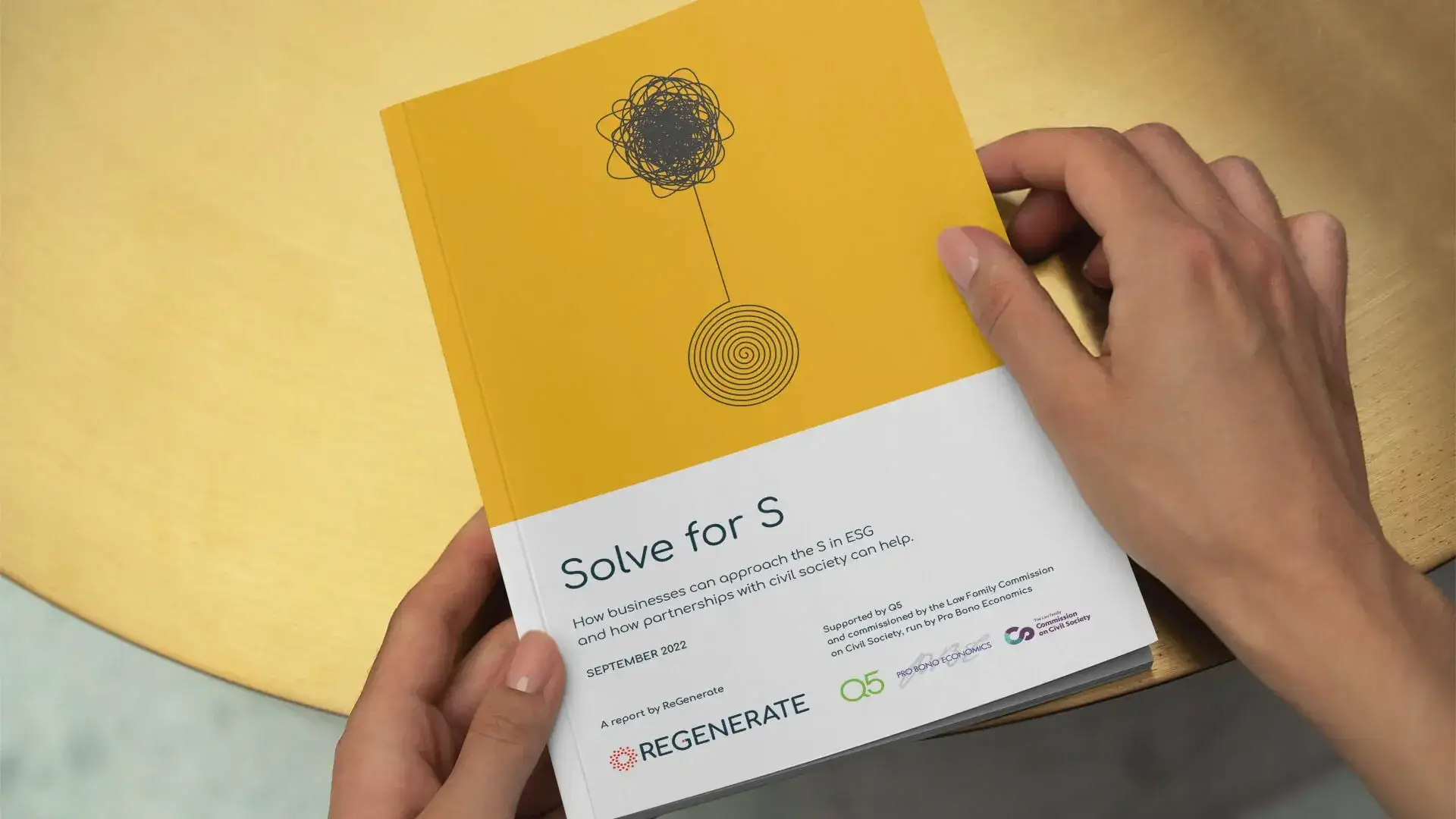Two hands holding ReGenerate's Solve for S report: a yellow cover with a fine line illustration above the title panel