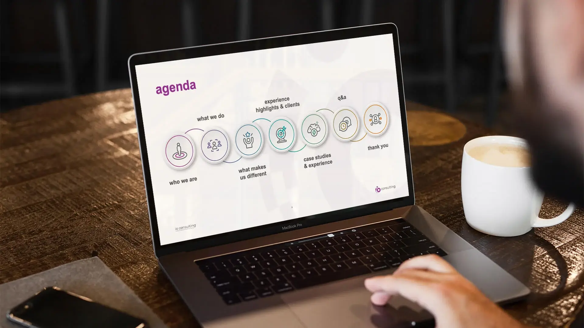A laptop on a desk showing the agenda slide of an io credentials deck, each section marked with a line drawn icon