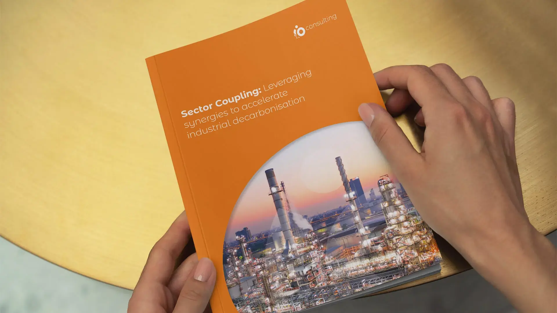 Hands holding io's Sector Coupling report, an orange cover with the io logo and a photograph of an industrial plant at dusk